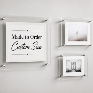 May include: Three clear acrylic floating frames with silver metal standoffs. The largest frame displays the text Made to Order Custom Size. The other two frames contain black and white photographs of a skyscraper in fog and a suspension bridge.