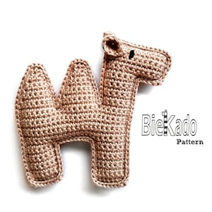 Puede incluir: Un juguete de crochet en forma de camello de color marrón claro. El juguete está hecho con un patrón de punto simple y tiene un pequeño ojo negro. El juguete está sentado sobre un fondo blanco. BieKado Pattern está escrito en texto negro en el lado derecho de la imagen.