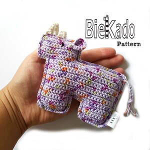 Puede incluir: Un juguete de animal de crochet de color morado y naranja con forma de rinoceronte con un cuerno blanco. El juguete está siendo sostenido en la mano de una persona. El texto "Biekado Pattern" es visible en el fondo.