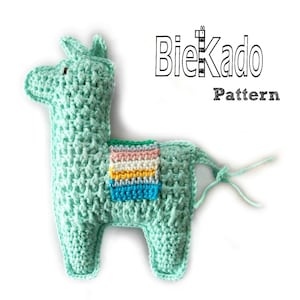 Puede incluir: Un juguete de llama de crochet verde con una franja de colores arcoíris en la espalda. La llama tiene un pequeño ojo negro y una nariz blanca. El juguete tiene una cola de cuerda.