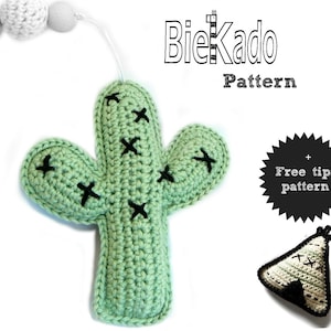 May include: A green crocheted cactus toy with black stitches. The cactus is hanging from a white string with white beads. The text "BieKado Pattern" is written in black above the cactus. A black and white circle with the text "Free tipi pattern" is in the bottom right corner of the image. A small crocheted tipi toy is below the circle.