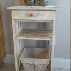May include: A white wooden side table with a drawer and two shelves. The top shelf has a collection of ceramic teapots and mugs. The bottom shelf has a wire basket with a fabric liner.