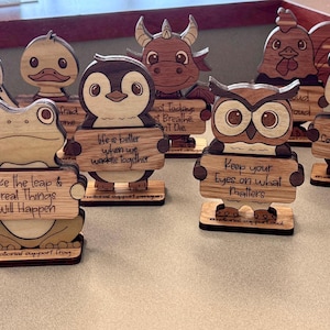 May include: Wooden animal figurines, including a unicorn, raccoon, duck, penguin, bull, rooster, owl, panda, and turtle, each holding a sign with an inspirational message. The figurines are made of light-colored wood.