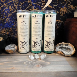 May include: Three white pillar candles with green labels. The labels read "Yule" and feature a black and white graphic design. The candles are on a wooden surface with crystals and a black cauldron.
