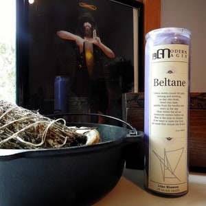 May include: Purple soy candle with a white label that reads "Modern Agix Beltane" and includes a description of the holiday. The candle is 18 oz and has a lilac blossom scent.