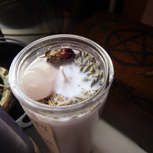 May include: A white candle with dried lavender, a rose bud, and a pink crystal inside a clear glass jar. The candle is on a wooden surface with a pentagram design.