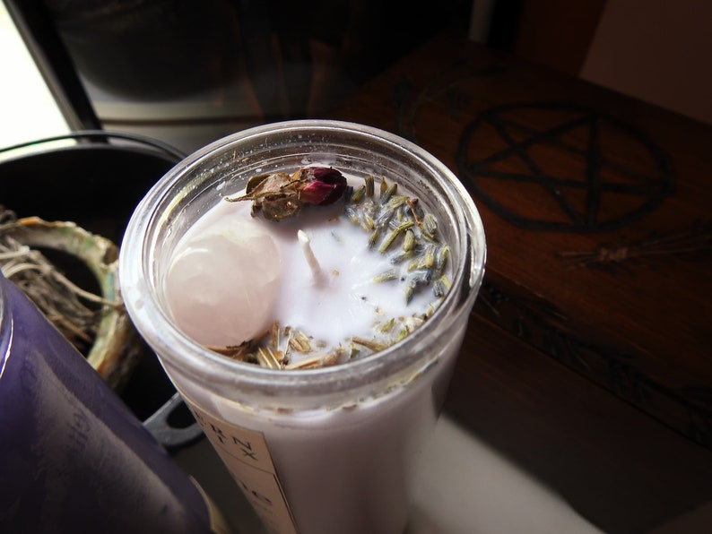 May include: A white candle with dried lavender, a rose bud, and a pink crystal inside a clear glass jar. The candle is on a wooden surface with a pentagram design.