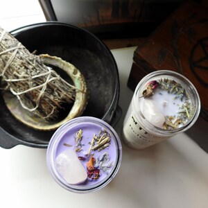 May include: Two purple glass candle jars with white wax and various dried herbs and flowers, including a rose and a clear quartz crystal. The candles are on a white surface.