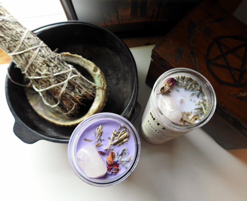 May include: Two purple glass candle jars with white wax and various dried herbs and flowers, including a rose and a clear quartz crystal. The candles are on a white surface.