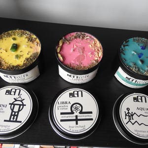 May include: Three scented soy candles in black tins with lids. The candles are yellow, pink, and teal, each with decorative elements and crystals. The lids feature zodiac symbols and candle scents: Champagne, Pear Orchard, and Mountain Mist. Each candle is 8 oz.