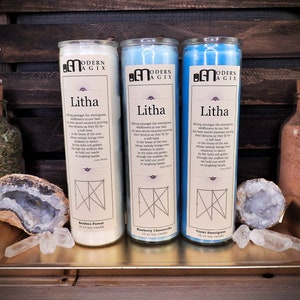 May include: Three white, blue, and light blue candles with the text "Litha" and "Modern Agix" on the labels. The candles are arranged on a gold tray with crystals and a jar of brown sand.
