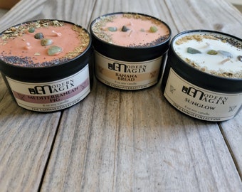May include: Three 8 ounce soy candles in black tins with labels that read "Modern Agix" and the scent names: "Mediterranean Fig", "Banana Bread", and "Sunglow". The candles are topped with dried herbs and crystals.