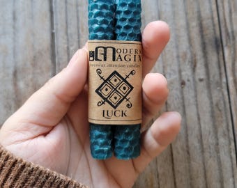 May include: Two teal beeswax intention candles wrapped with a brown paper label. The label reads "MODERN MAGIX beeswax intention candles" with a geometric design and the word "LUCK". The candles have a honeycomb texture.