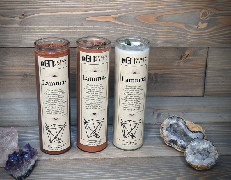 May include: Three glass candles with a brown, orange, and white exterior. Each candle has a label that reads "Modern Agix" and "Lammas". The candles are arranged in a row on a wooden surface. The candles are surrounded by crystals.
