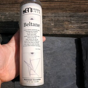 May include: A white soy candle with a label that reads "Beltane" and a poem about the celebration. The candle is 16 oz and has a watermint scent. The candle is sitting on a wooden surface.