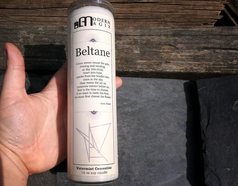 May include: A white soy candle with a label that reads "Beltane" and a poem about the celebration. The candle is 16 oz and has a watermint scent. The candle is sitting on a wooden surface.