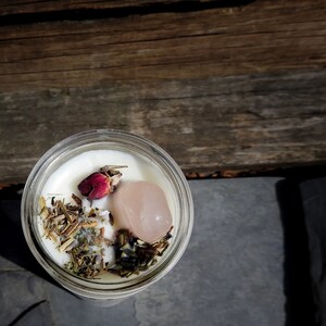 May include: A white candle with dried herbs, a pink rose bud, and a pink crystal inside a clear glass jar.