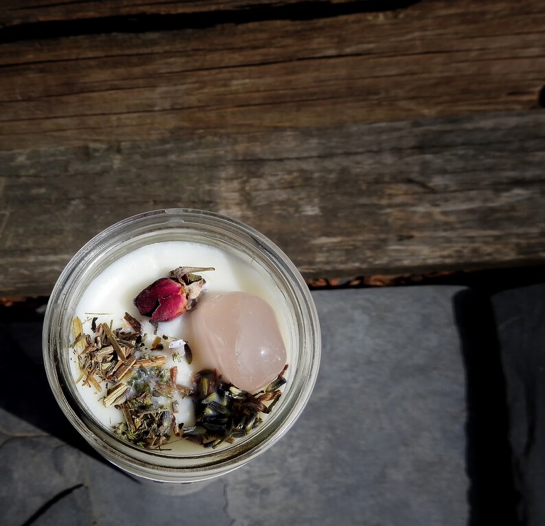 May include: A white candle with dried herbs, a pink rose bud, and a pink crystal inside a clear glass jar.