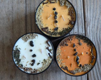 May include: Three round candles with black and orange wax and various dried herbs and spices on top. The candles are in black containers.