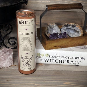 May include: A brown soy candle with the text "Lammas" and a description of the holiday. The candle is labeled "Modern Magick" and is 10 oz. The candle is sitting on a wooden surface with a book titled "The Element Encyclopedia of Witchcraft" and a wooden box with amethyst crystals.