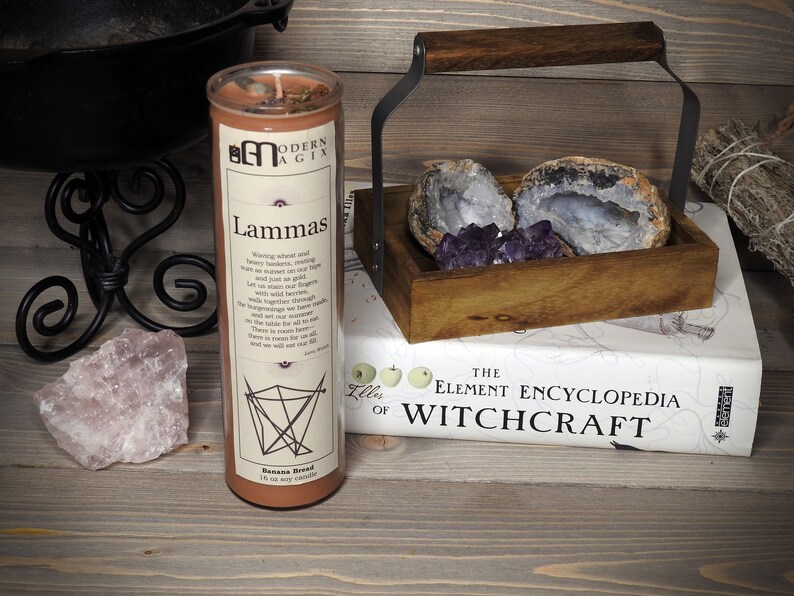 May include: A brown soy candle with the text "Lammas" and a description of the holiday. The candle is labeled "Modern Magick" and is 10 oz. The candle is sitting on a wooden surface with a book titled "The Element Encyclopedia of Witchcraft" and a wooden box with amethyst crystals.