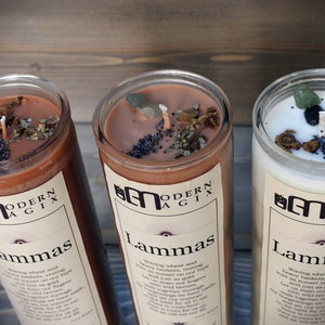 May include: Three brown glass candles with a white label that reads "Modern Agix" and "Lammas". The candles are topped with dried herbs, crystals, and black poppy seeds.