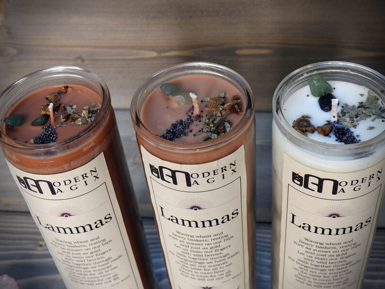 May include: Three brown glass candles with a white label that reads "Modern Agix" and "Lammas". The candles are topped with dried herbs, crystals, and black poppy seeds.