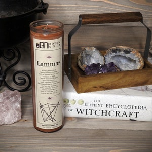 May include: A brown candle with a label that reads "Modern Agix" and "Lammas". The label also includes a poem about the harvest festival. The candle is sitting on a wooden surface with a book titled "The Element Encyclopedia of Witchcraft" and a wooden box with crystals.