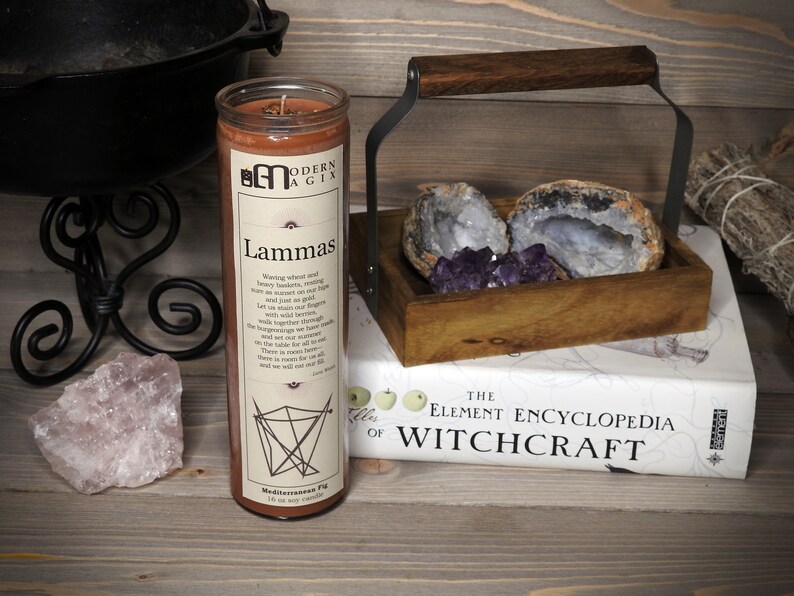 May include: A brown candle with a label that reads "Modern Agix" and "Lammas". The label also includes a poem about the harvest festival. The candle is sitting on a wooden surface with a book titled "The Element Encyclopedia of Witchcraft" and a wooden box with crystals.