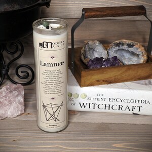 May include: A white candle with a black and white label that reads "Lammas" and a description of the holiday. The candle is sitting on a wooden surface with a book titled "The Element Encyclopedia of Witchcraft" and a wooden box with amethyst crystals and agate.