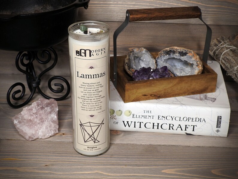 May include: A white candle with a black and white label that reads "Lammas" and a description of the holiday. The candle is sitting on a wooden surface with a book titled "The Element Encyclopedia of Witchcraft" and a wooden box with amethyst crystals and agate.