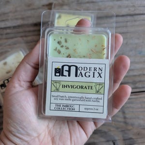 Puede incluir: Un peque&ntilde;o recipiente de pl&aacute;stico transparente contiene una fusi&oacute;n de cera de soja verde con motas doradas. La etiqueta dice "MODERN AGIX" e "INVIGORATE". El texto debajo dice "Lote peque&ntilde;o, hecho a mano intencionalmente, fusiones de cera de soja adornadas con hierbas. THE IMBOLC COLLECTION aprox. 2 oz."