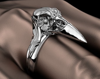 May include: A silver ring with a detailed raven skull design. The ring's band has intricate floral and leaf patterns. The raven's beak extends outwards, and the skull is adorned with a flower. The ring is set against a brown background.