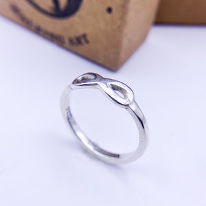 Puede incluir: Un anillo plateado con un diseño de símbolo de infinito. El anillo está sobre un fondo blanco, con una caja marrón borrosa en el fondo. El símbolo de infinito es el punto focal del anillo.