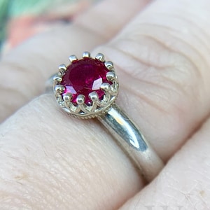 Può includere: Un anello d'argento con una pietra preziosa di rubino rosso incastonata in una montatura a forma di corona.