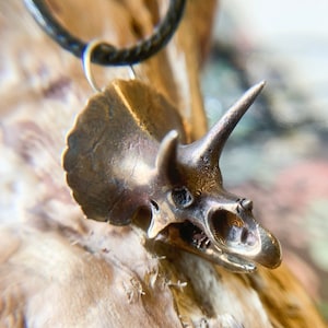 May include: A bronze-colored pendant necklace featuring a detailed triceratops skull. The pendant is suspended from a black cord. The dinosaur skull has a prominent horn and a frill around the back of the head. The necklace is displayed on a wooden surface.