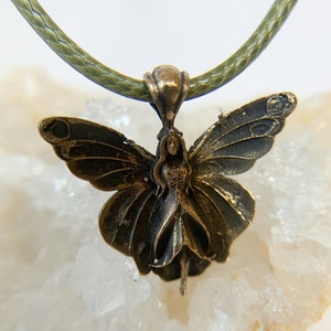 Puede incluir: Un collar con colgante de color bronce que representa un hada con alas de mariposa. El hada está centrada dentro de las alas, y el collar tiene un cordón verde. El colgante es detallado y parece ser de metal.