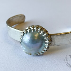 May include: A silver cuff bracelet featuring a large, iridescent, round pearl. The pearl is set in a decorative silver bezel. The bracelet has a simple, brushed metal band. The overall design is elegant and minimalist.