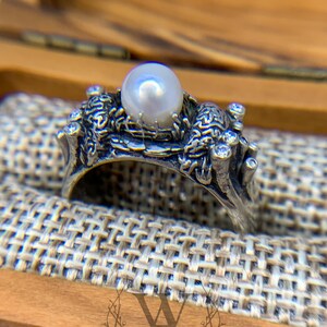 May include: A silver ring with a pearl and intricate detailing. The ring is set on a textured fabric surface.