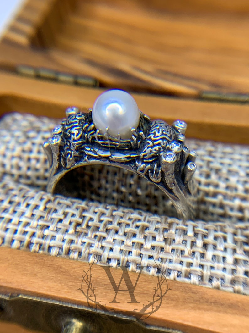 May include: A silver ring with a pearl and intricate detailing. The ring is set on a textured fabric surface.