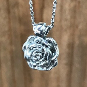 Puede incluir: Collar con colgante plateado con forma de rosa. El colgante muestra una rosa detallada, con pétalos individuales visibles. El collar cuelga de una fina cadena. El fondo es borroso y de tono neutro.