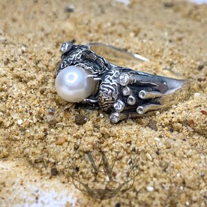 May include: A silver ring with a large white pearl and smaller clear stones set in a detailed design. The ring is on a sandy surface.