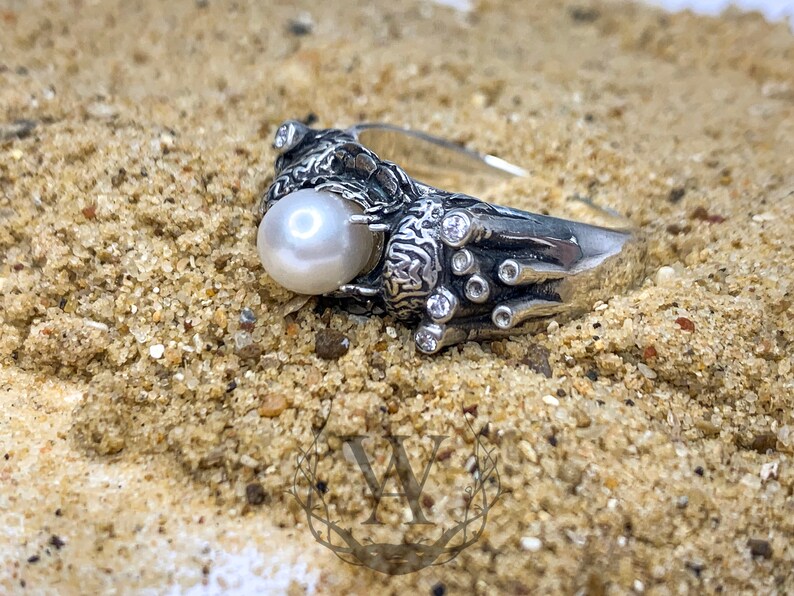 May include: A silver ring with a large white pearl and smaller clear stones set in a detailed design. The ring is on a sandy surface.