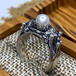 May include: A silver ring with a pearl set in a detailed design of branches and leaves. The ring is in a wooden box with a fabric lining.