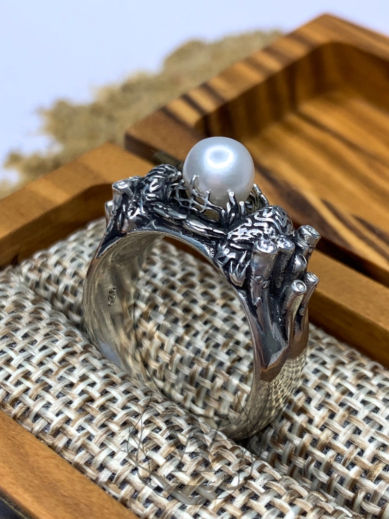 May include: A silver ring with a pearl set in a detailed design of branches and leaves. The ring is in a wooden box with a fabric lining.