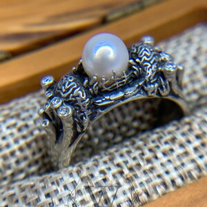 May include: A silver ring with a pearl set in a detailed design of branches and leaves.
