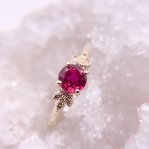 May include: A gold ring featuring a round, vibrant red gemstone. The ring has delicate leaf-like designs on either side of the gemstone. The band is thin and polished, creating an elegant and refined aesthetic. The ring is set against a soft, blurred background.