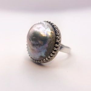 May include: A silver ring featuring a large, iridescent gray pearl. The pearl has a unique, irregular shape and is set in a decorative silver bezel with a beaded edge. The ring's band is a simple, polished silver.