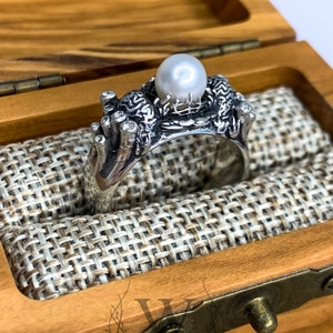 May include: A silver ring with a pearl and intricate design, displayed in a wooden box with a fabric lining.