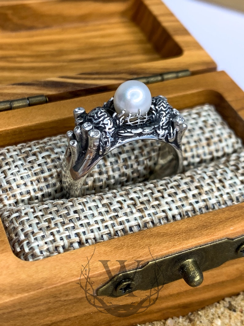 May include: A silver ring with a pearl and intricate design, displayed in a wooden box with a fabric lining.